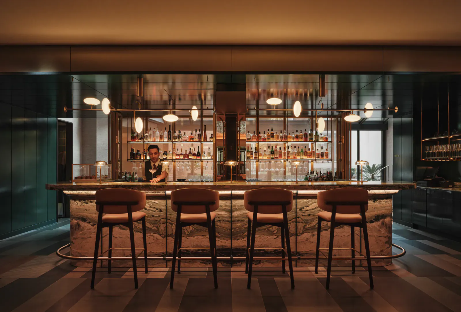 Elegant marble bar with four upholstered stools, bartender preparing drinks behind illuminated shelves of bottles