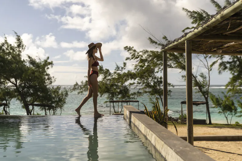Woman in bikini and hat walking along infinity pool edge overlooking turquoise ocean and palm trees at Constance Tekoma, Mauritius