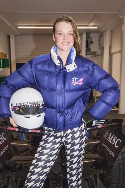 Young woman in blue puffer jacket with Alpine 4 logo holding white racing helmet, standing beside F1 car in garage