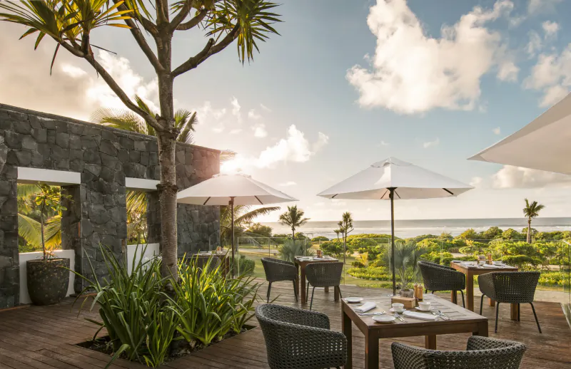Luxury oceanfront restaurant terrace with wooden tables, woven chairs, white umbrellas, and tropical plants overlooking the sea at sunset