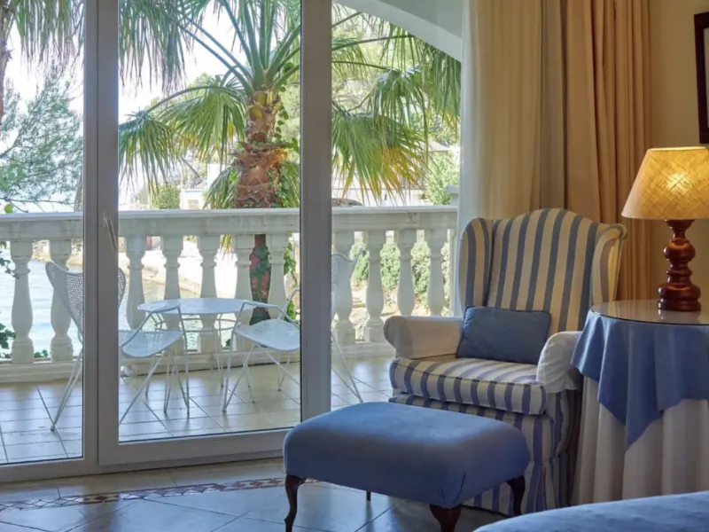 Bright hotel room with striped armchair and blue ottoman beside glass doors opening to a sunny balcony with palm trees and white patio furniture