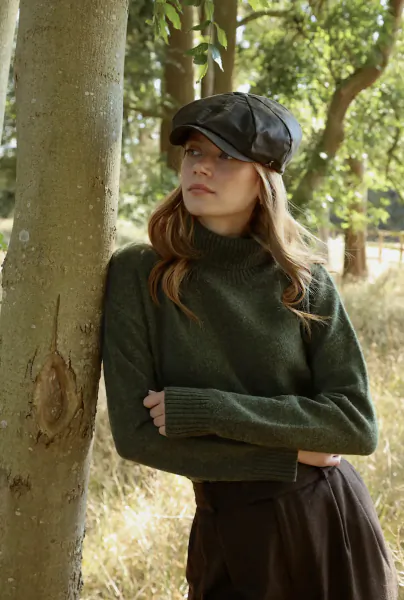 Woman in green turtleneck sweater and black leather cap leaning against tree in forest