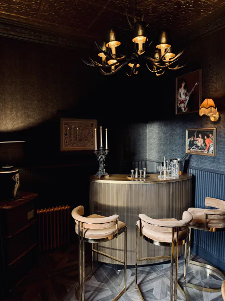 Elegant dark bar with curved metallic counter, three beige barstools, antler chandelier, and framed artwork on textured walls