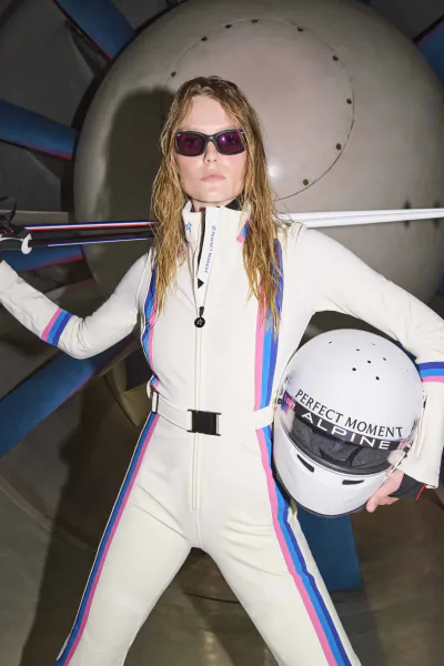 Woman in white racing suit with blue and pink stripes holding helmet, standing in front of jet engine wearing sunglasses