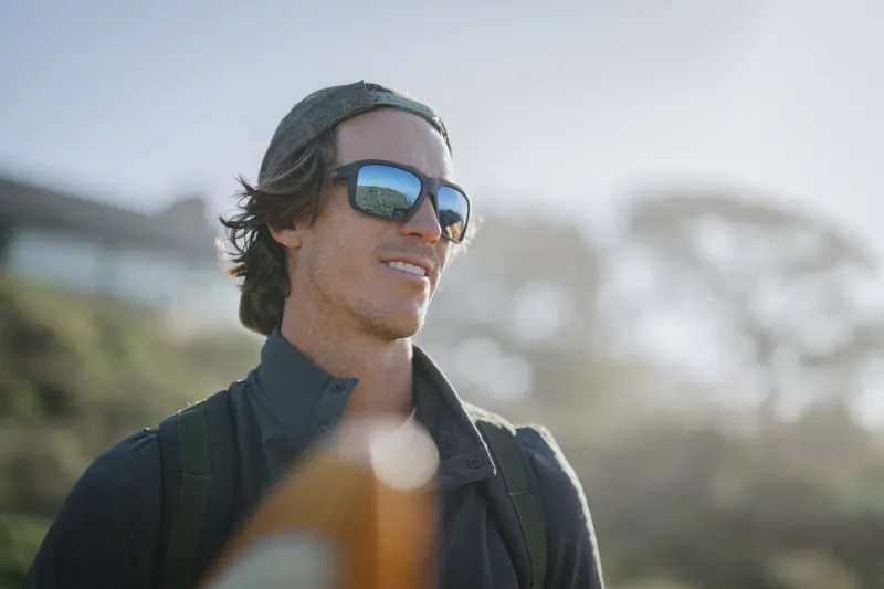 Smiling man wearing SMITH sunglasses and cap with backpack outdoors in sunlight