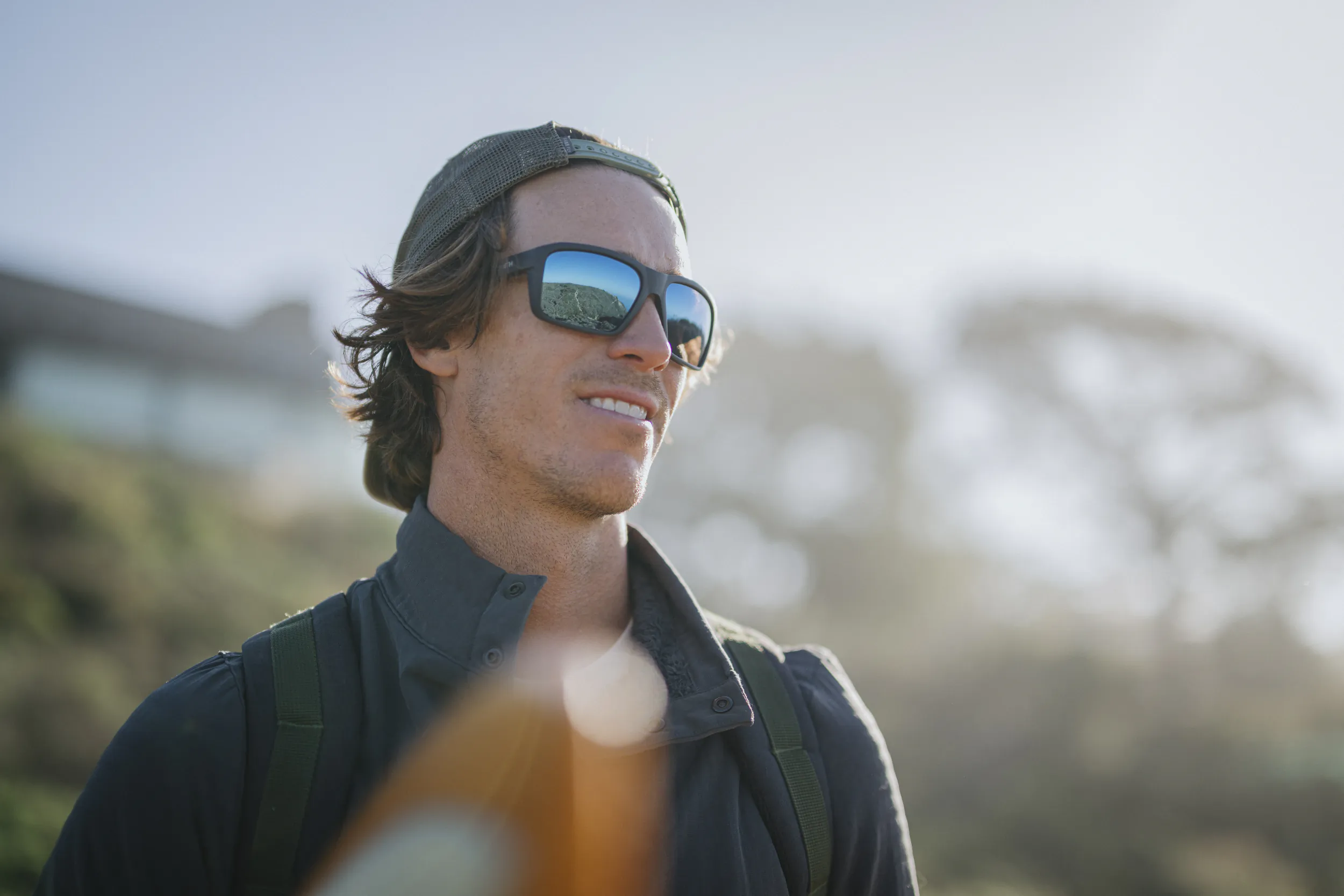Smiling man wearing SMITH sunglasses and cap with backpack outdoors in sunlight