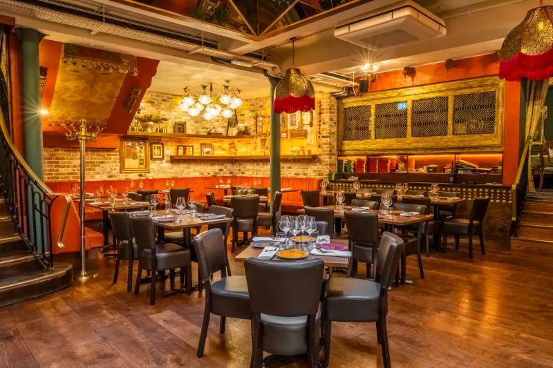 Elegant Indian restaurant interior with dark leather chairs, wooden tables set with wine glasses, exposed brick walls, and ornate golden lamps