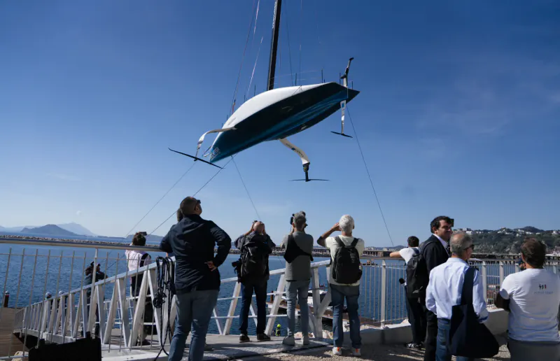 Spectators watch a blue and white AC75 sailboat being lifted by crane over a harbor dock