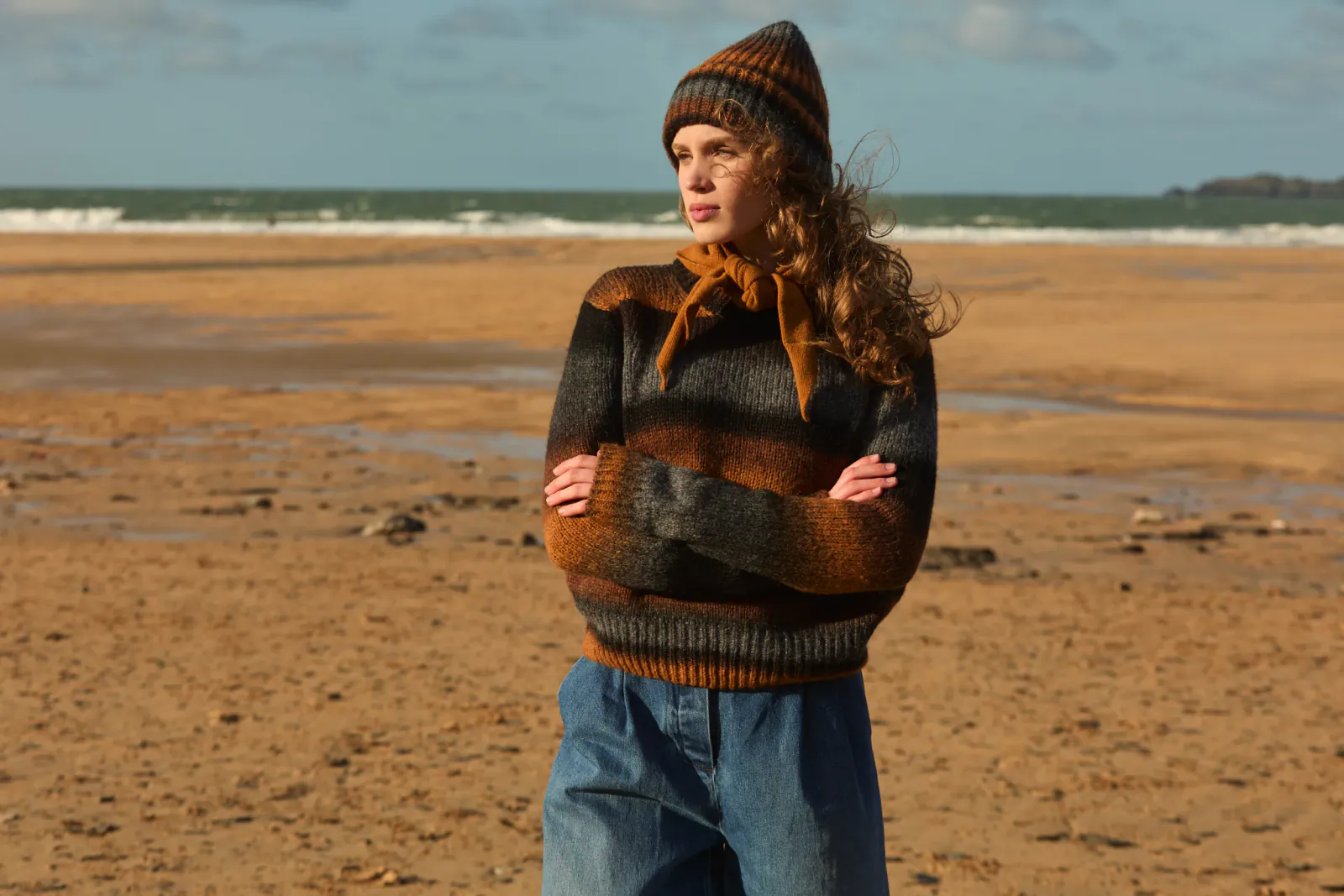 Young woman with curly red hair wearing striped knit sweater, beanie and jeans standing with arms crossed on a sandy beach