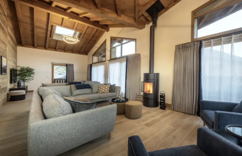 Modern open-plan living room with vaulted wood ceiling, skylights, grey sectional sofa, lit wood-burning stove, and large windows overlooking mountains