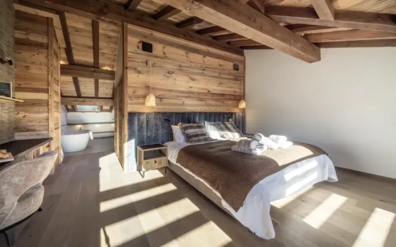 Rustic wooden bedroom with large bed, exposed beams, and a freestanding bathtub in the background