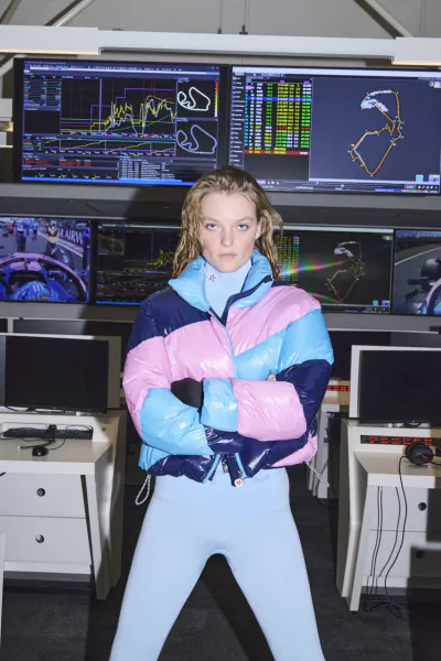 Woman in pink and blue puffer jacket stands with arms crossed in front of F1 racing data screens