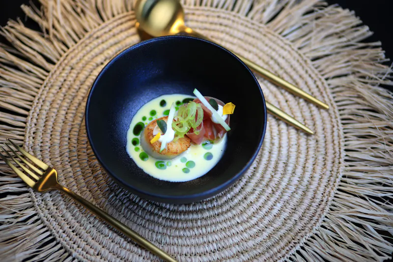 Seared scallop in black bowl with green sauce, microgreens, and gold cutlery on woven placemat