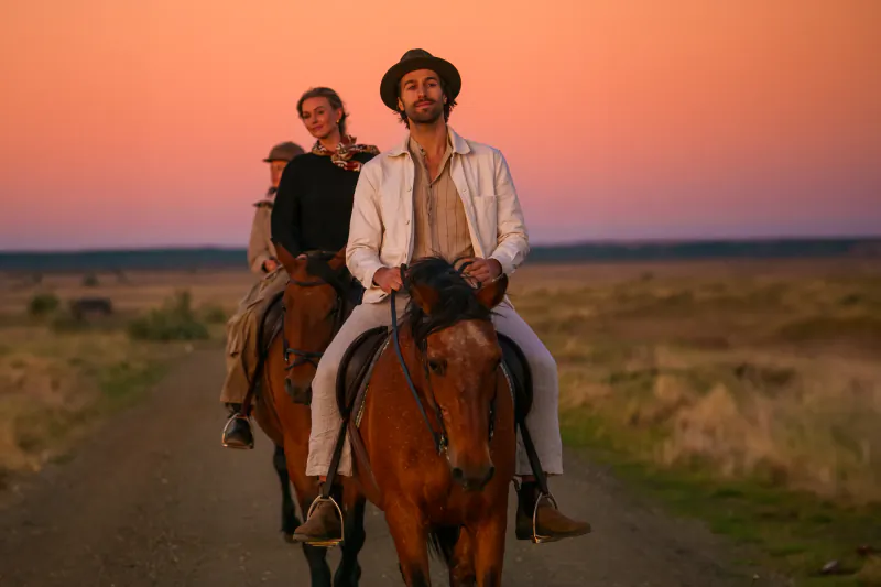 Three people riding horses on a dirt road at sunset, man in white shirt and hat leading in front