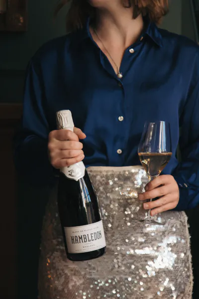 Woman in blue shirt and sequin skirt holding Hambledon sparkling wine bottle and glass of white wine
