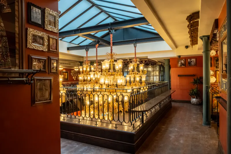 Elegant restaurant interior with ornate golden chandeliers above a central dining area, framed pictures on red walls, and a skylight ceiling