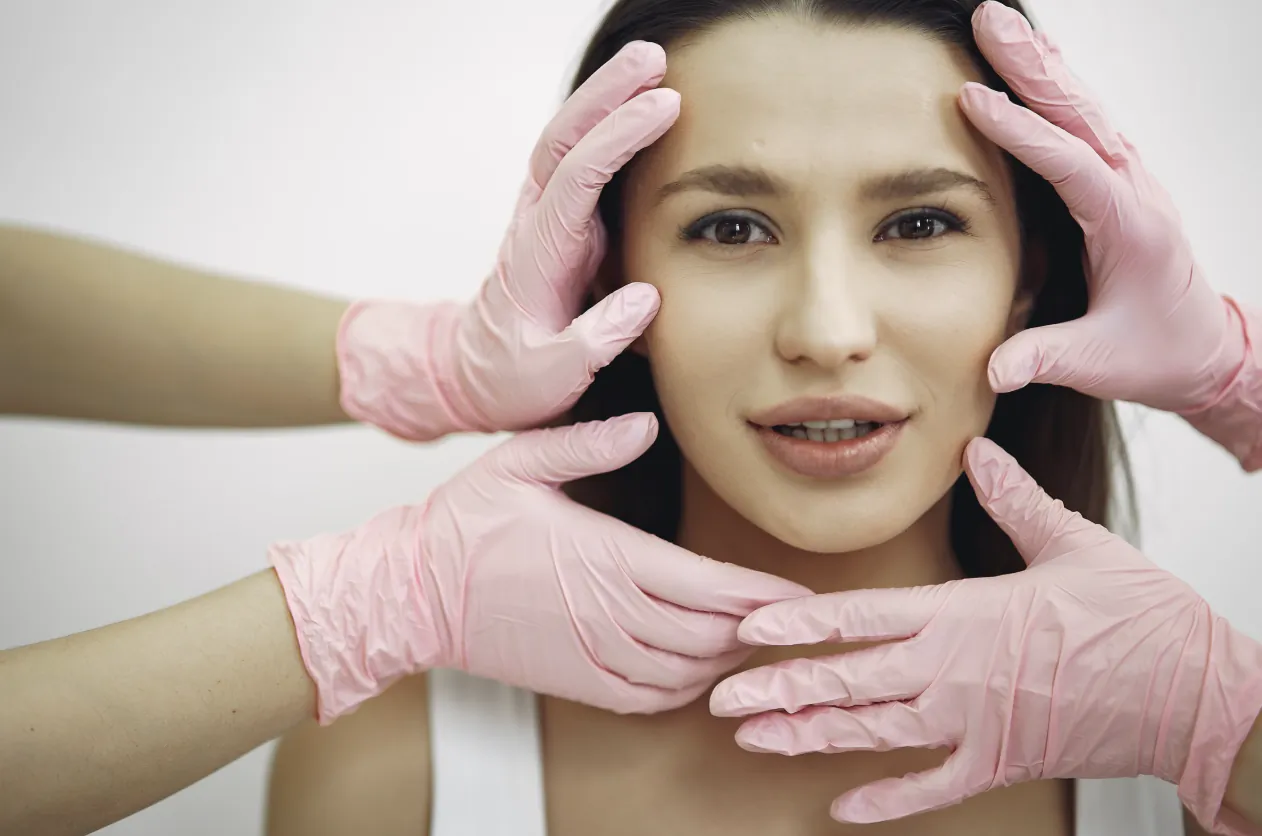 Woman receiving facial assessment from gloved hands examining her forehead, cheeks and jawline
