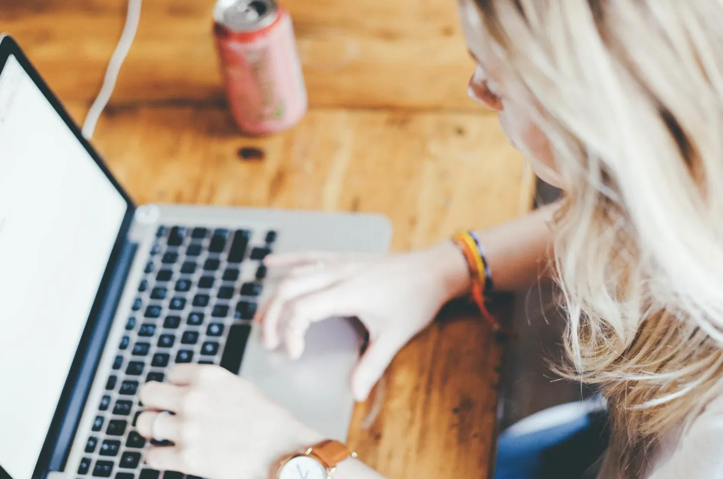 Woman typing on laptop at wooden table with pink soda can nearby