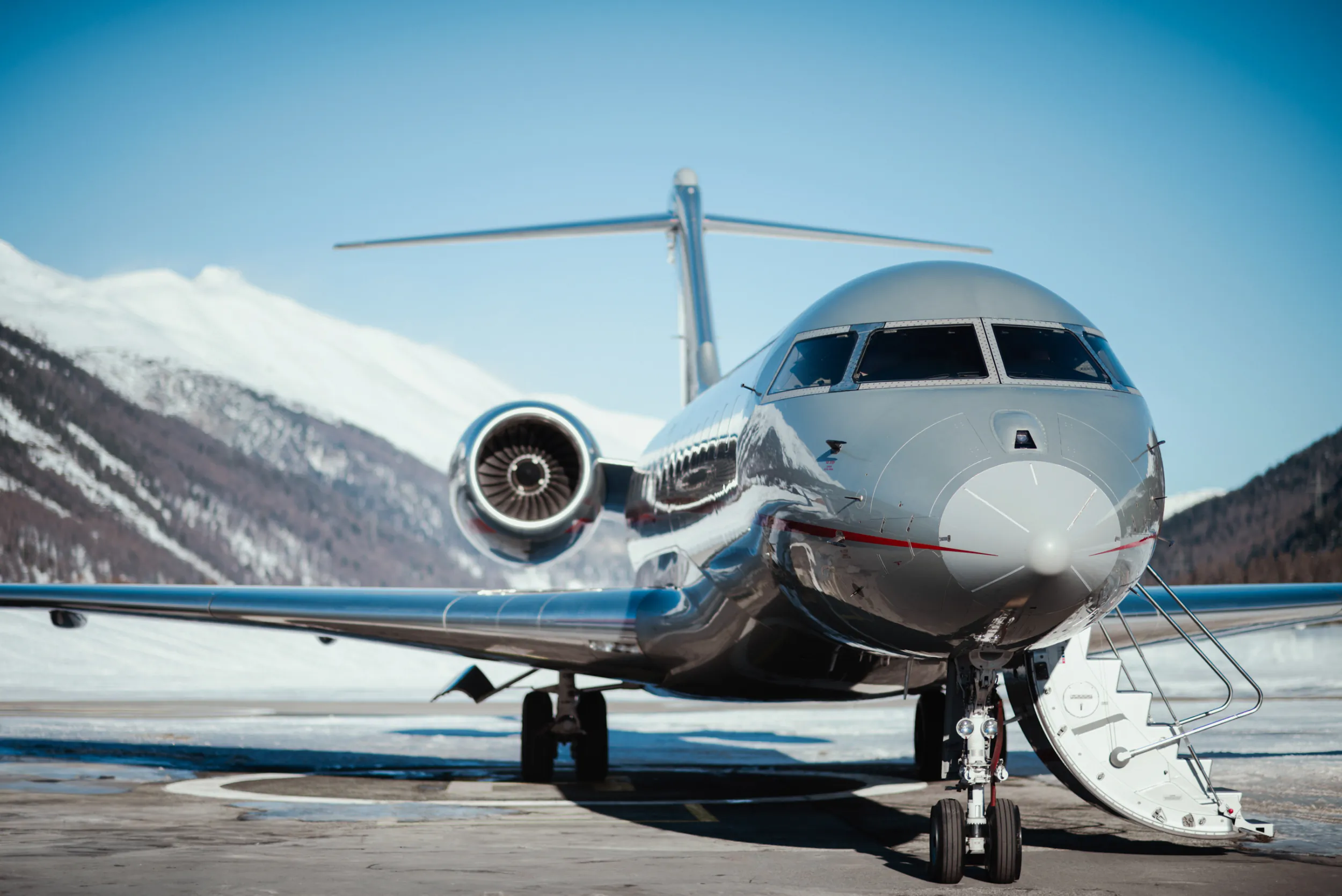 Silver private jet parked on snowy tarmac with mountains and blue sky in background
