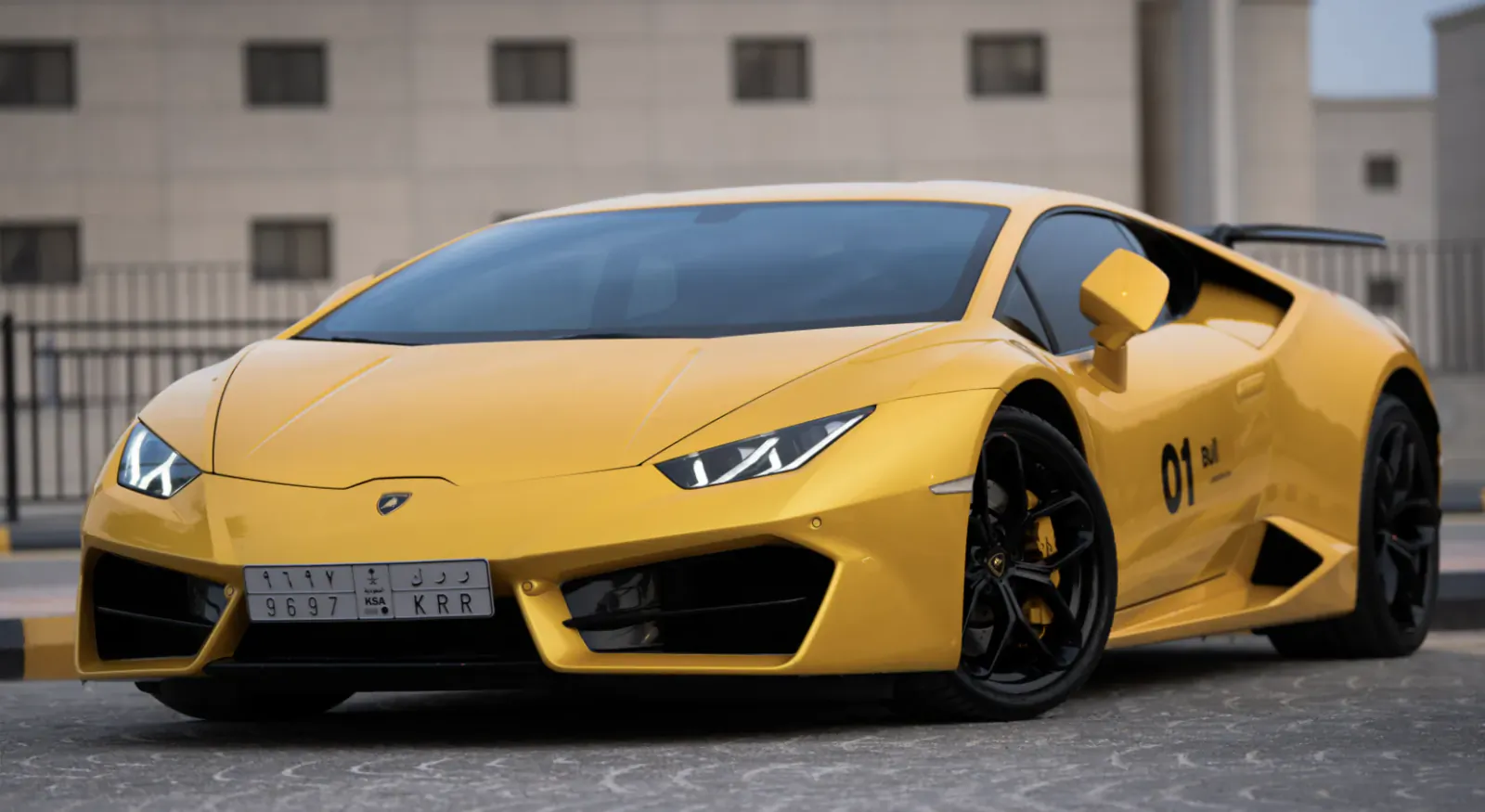 Yellow Lamborghini Huracan sports car with black wheels and '01' decal parked in front of a modern building
