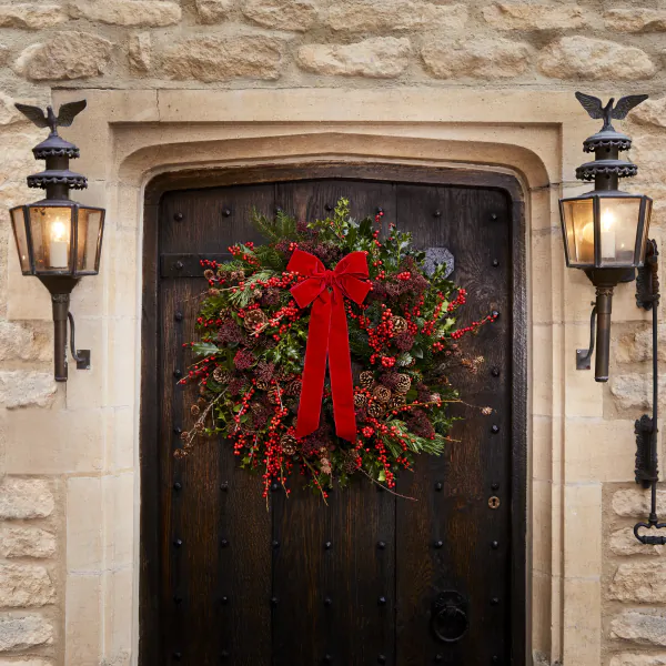 Christmas wreath with red bow and berries on dark wooden door flanked by lit lanterns