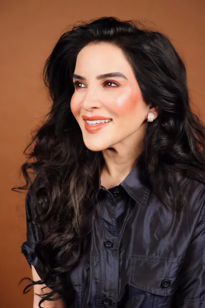 Dr. Vali smiling with long dark wavy hair, wearing a dark button-up shirt against a warm brown backdrop