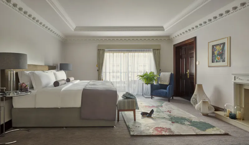 Luxury hotel bedroom with large bed, white linens, blue armchair, fireplace, and floral rug in an elegant London suite