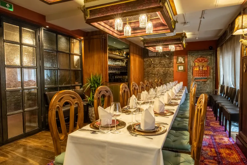 Elegant restaurant dining room with long white-clothed table set for a formal meal, ornate wooden chairs, and warm ambient lighting