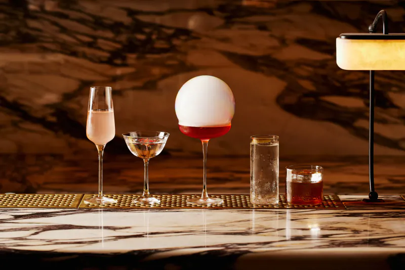 Four cocktails on a marble bar: champagne flute, coupe glass, large sphere with red liquid, and two short glasses beside a glowing lamp
