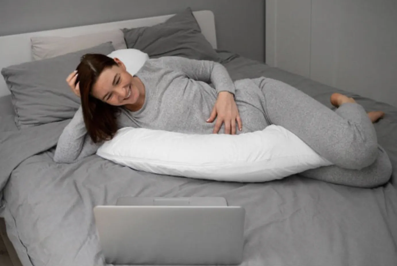 Pregnant woman in grey loungewear lying on bed with white pregnancy pillow, smiling while watching laptop