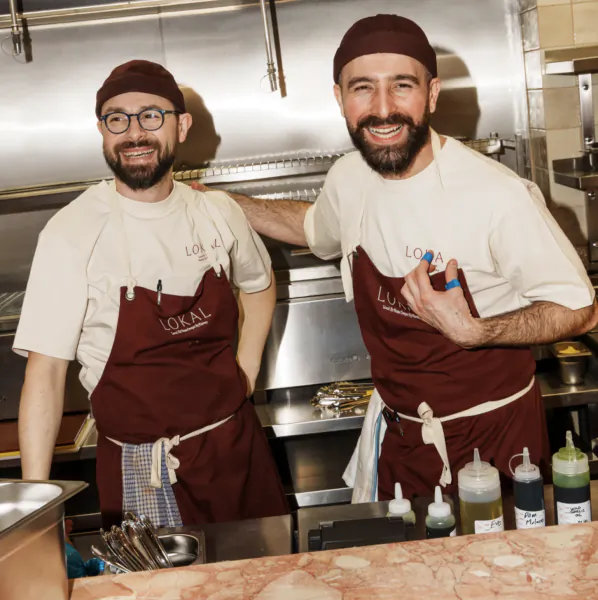 Two smiling male chefs in white shirts and maroon aprons stand in a professional kitchen, one with his arm around the other