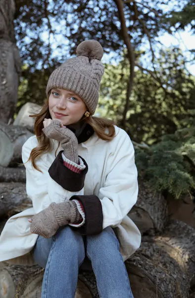 Young woman in beige pom-pom beanie and matching mittens sits on a log wearing a white coat and jeans