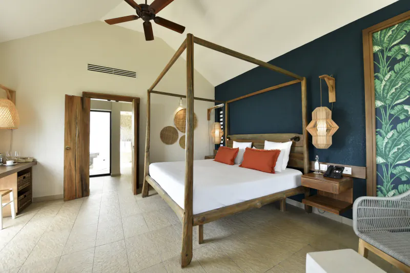 Luxury hotel bedroom with four-poster wooden bed, white linens and orange pillows, dark blue accent wall with tropical leaf wallpaper, ceiling fan, and open door to balcony