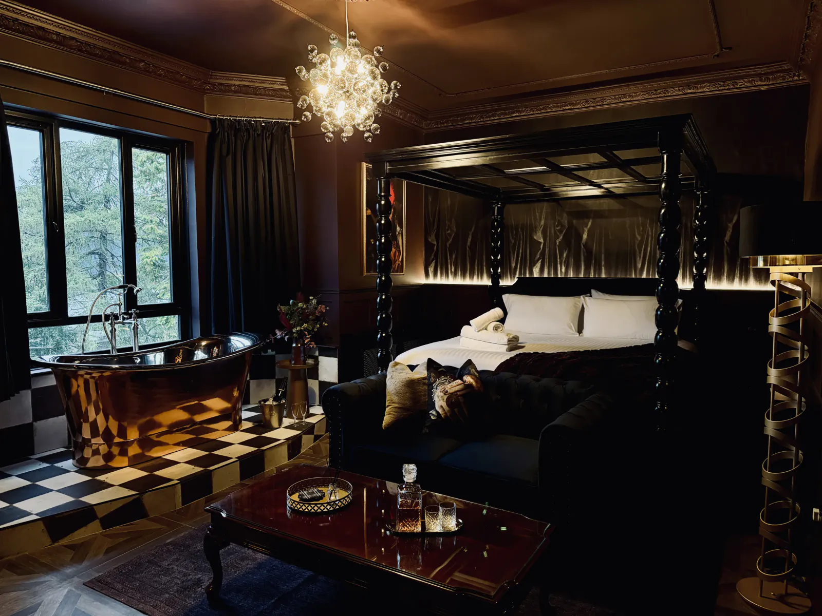 Luxurious dark bedroom with four-poster bed, copper bathtub by window, black sofa with dog, and ornate chandelier
