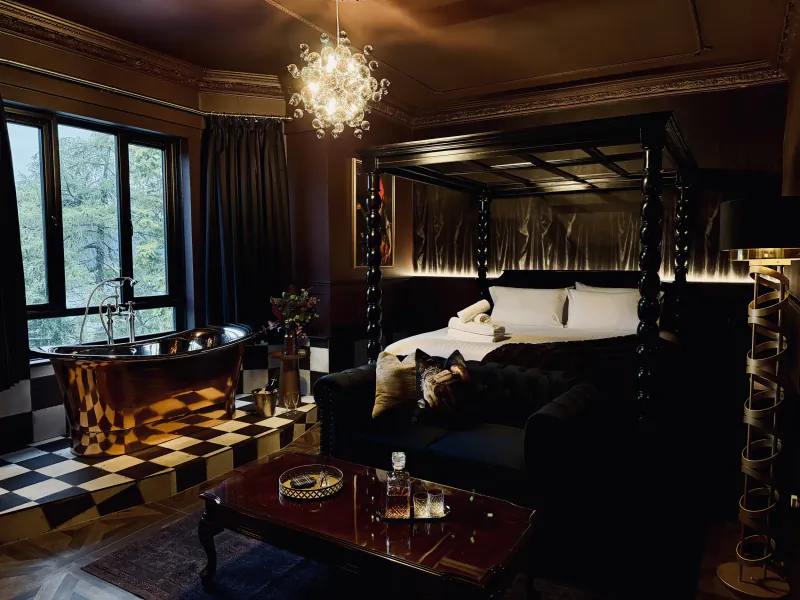 Luxurious dark bedroom with four-poster bed, copper bathtub by window, black sofa with dog, and ornate chandelier