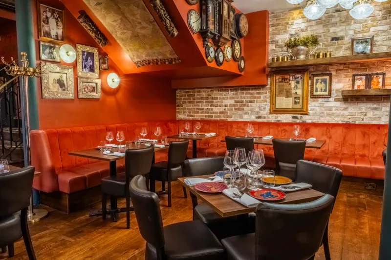 Elegant Indian restaurant interior with red leather banquettes, dark wood tables set for dining, exposed brick walls, and vintage clocks