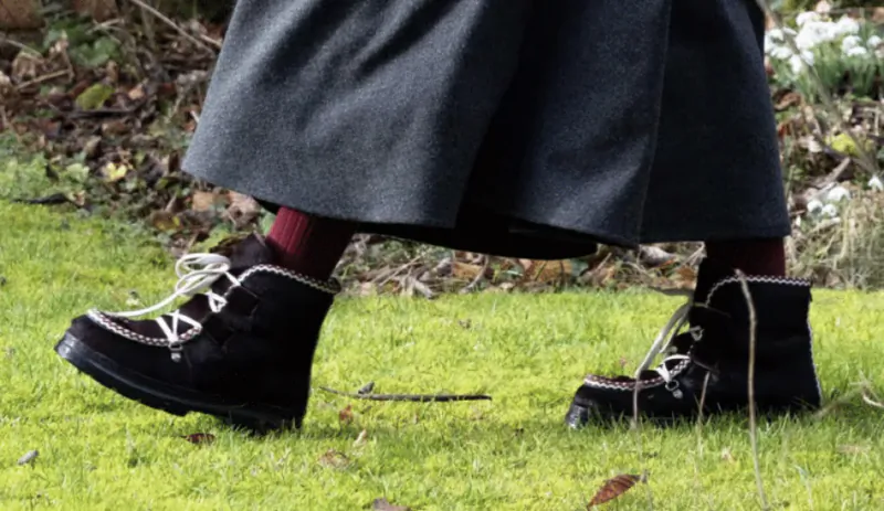 Close-up of black suede ankle boots with white laces and silver chain detail, worn with maroon socks and a grey skirt on grass