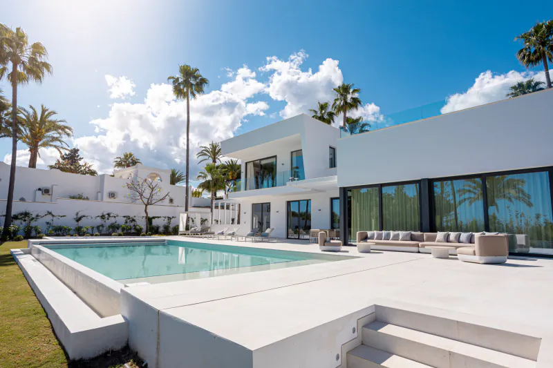 Modern white luxury villa with large pool, palm trees, and outdoor lounge area under bright blue sky