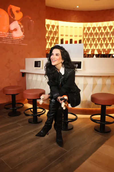 Woman in black outfit and boots posing on a bar stool in a modern orange and white bar with geometric wall patterns