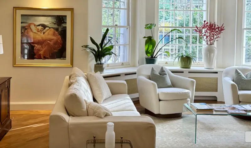Elegant living room with cream sofa, two white armchairs, glass coffee table, large framed classical painting, and potted plants by tall windows