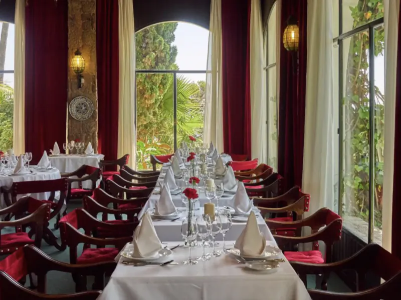 Elegant restaurant dining room with long white table set for formal dinner, red velvet chairs, arched windows overlooking garden