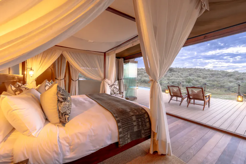 Luxury safari tent bedroom with white linens, canopy drapes, and wooden deck overlooking savanna landscape at dusk