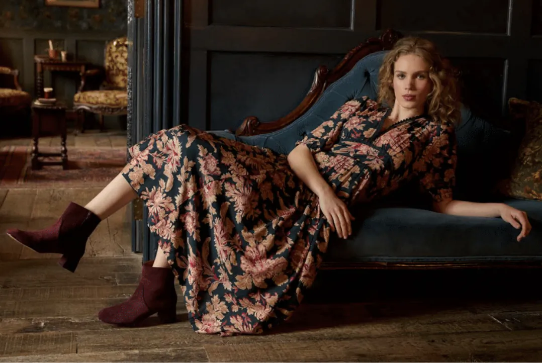 Woman in floral dress reclining on blue velvet chaise lounge wearing burgundy ankle boots