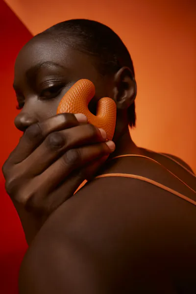 Close-up of a Black woman with short hair holding an orange textured micro-contouring tool against her cheek against a vibrant red-orange background