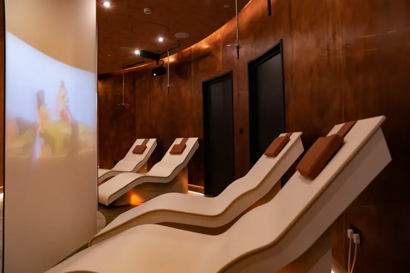 Modern spa relaxation room with four white ergonomic loungers, warm wood walls, and a large projection screen showing a sunset landscape