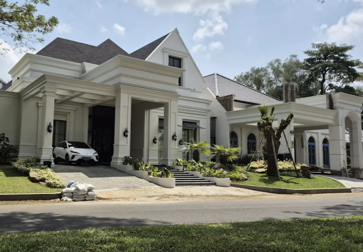 Modern white mansion with covered portico, white sports car parked in driveway, landscaped garden and paved road in foreground