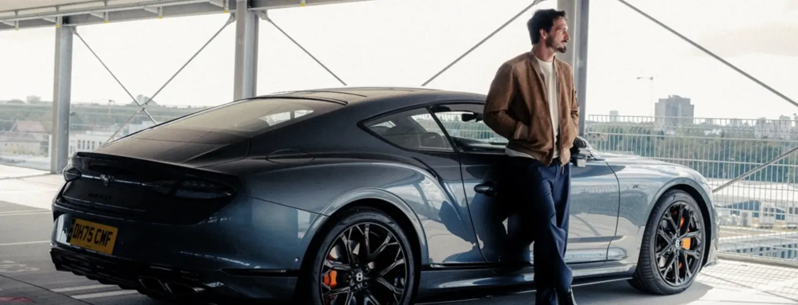 Man in brown jacket leaning against a dark blue Bentley sports car on a rooftop parking deck