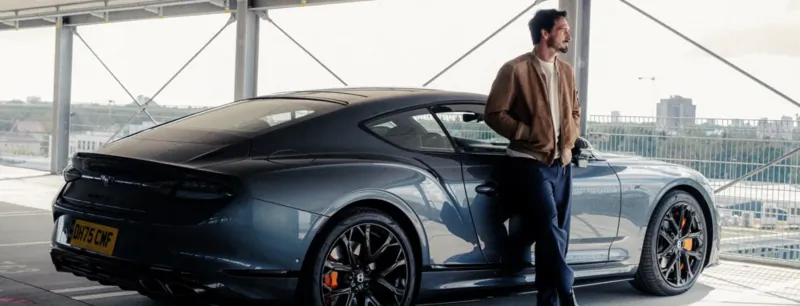 Man in brown jacket leaning against a dark blue Bentley sports car on a rooftop parking deck