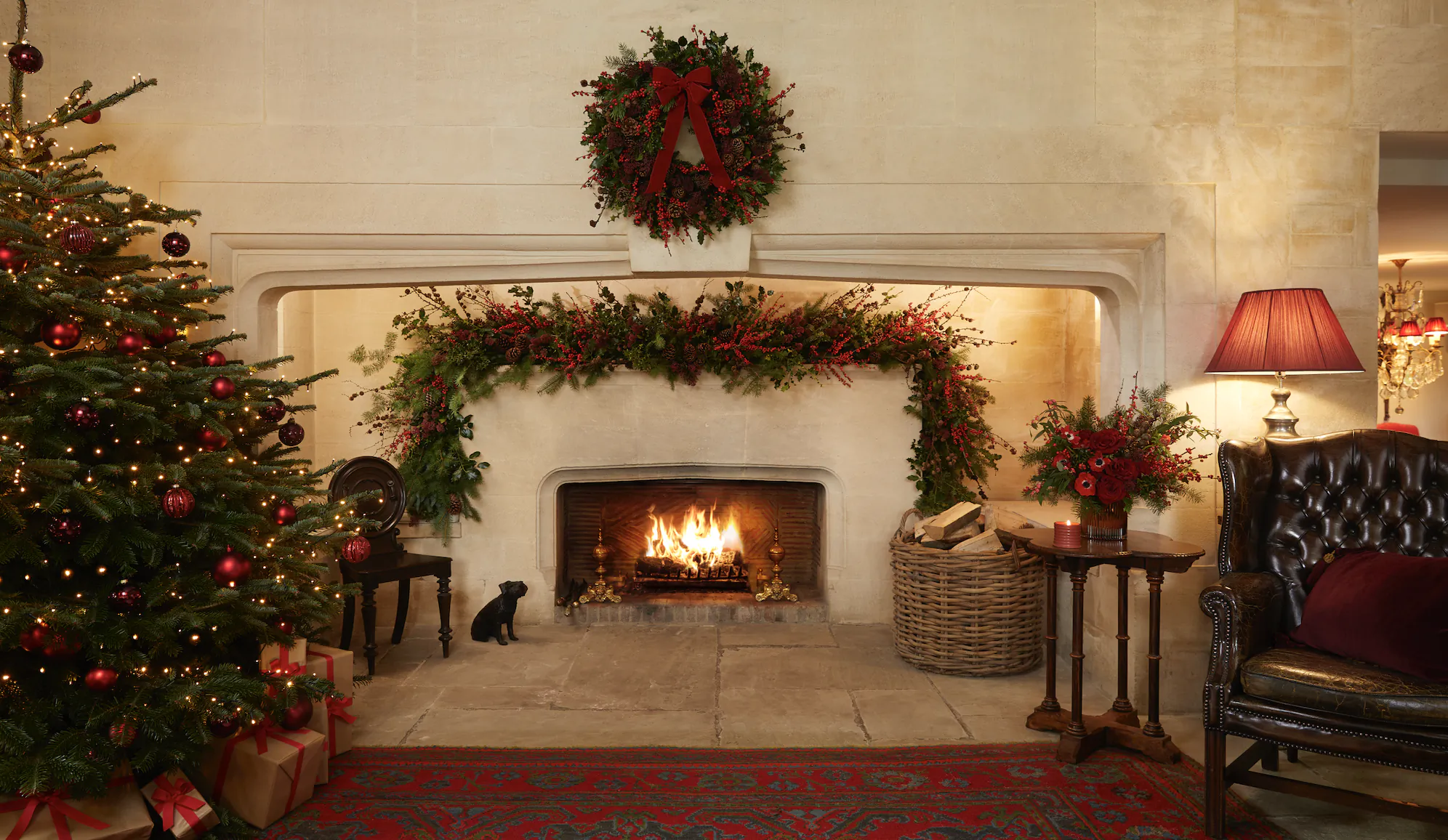 Festive living room with lit Christmas tree, roaring fireplace, red wreath above mantel, black cat sitting nearby, and leather armchair