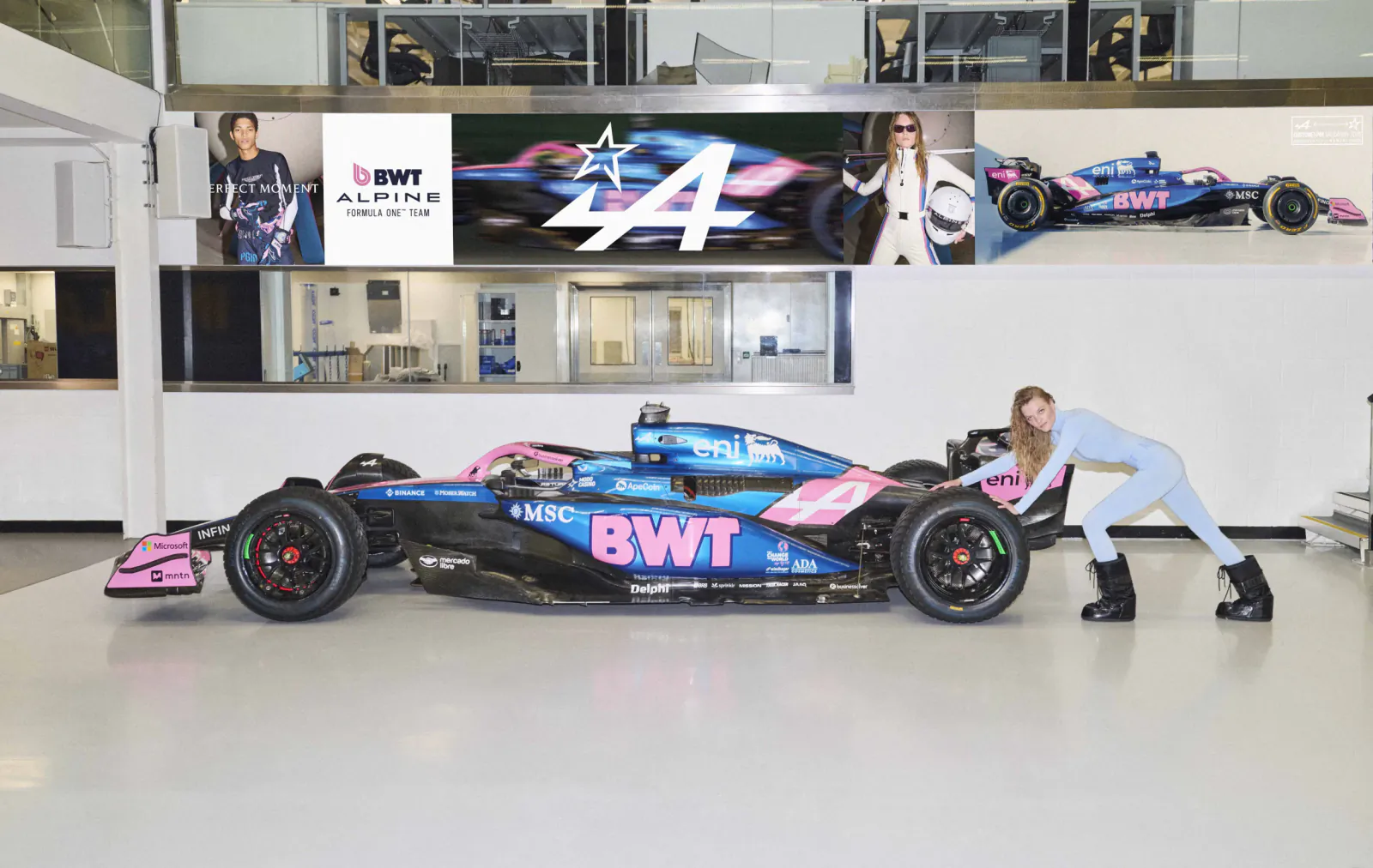 Woman in light blue outfit posing beside blue and pink BWT Alpine F1 race car in showroom